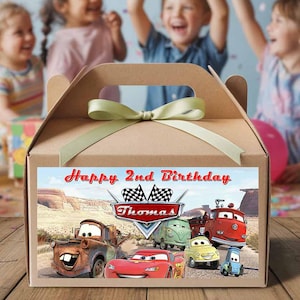 Może przedstawiać: Brązowe kartonowe pudełko na upominki z zieloną wstążką. Pudełko posiada kolorową etykietę z motywem Cars z napisem "Happy 2nd Birthday" i różnymi postaciami z kreskówek. Dzieci w tle.