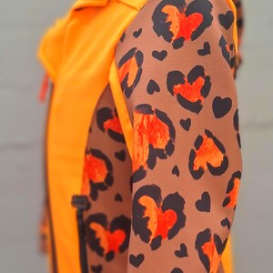 May include: Orange jacket with a brown and orange leopard print on the sleeves. The jacket has a zipper closure and a hood.