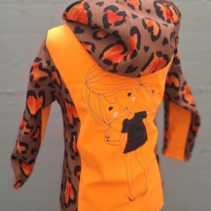May include: Orange and brown hooded jacket with a leopard print design. The jacket has a cute cartoon girl graphic on the back.
