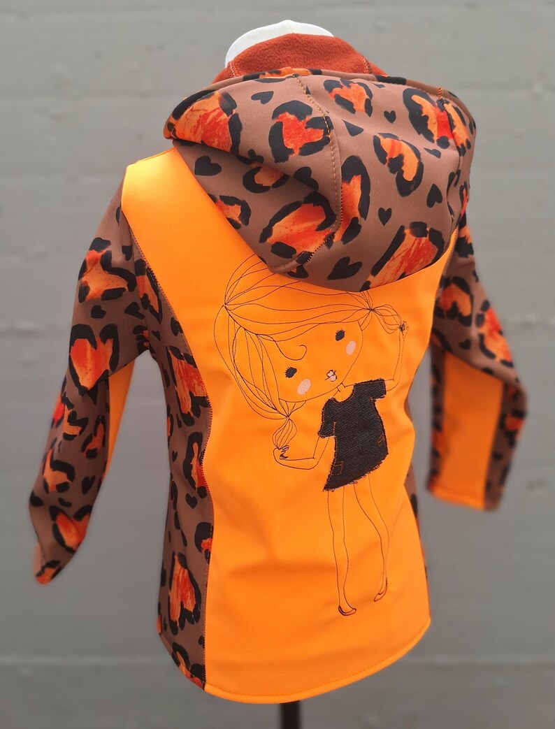 May include: Orange and brown hooded jacket with a leopard print design. The jacket has a cute cartoon girl graphic on the back.