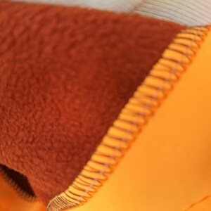 May include: Close-up of a brown fleece lining sewn to a bright orange fabric. The seam is visible and the fabric is textured.