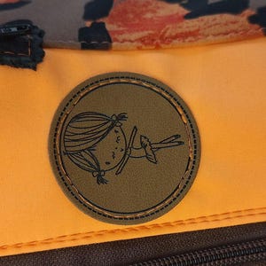 May include: Orange and brown backpack with a brown leather patch featuring a cartoon girl. The patch has a stitched border.