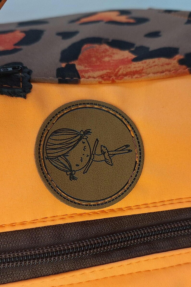 May include: Orange and brown backpack with a brown leather patch featuring a cartoon girl. The patch has a stitched border.