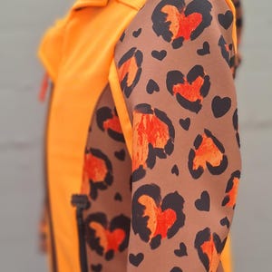 May include: Orange and brown jacket with a leopard print pattern on the sleeves. The jacket has a zip fastening and a hood.