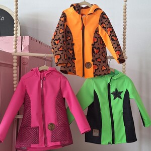 May include: Three children's jackets hanging on a wooden rack. The jackets are in different colours and patterns. One is orange with a brown leopard print, one is pink with a quilted pattern, and one is green with a black star.