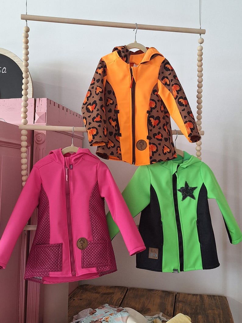 May include: Three children's jackets hanging on a wooden rack. The jackets are in different colours and patterns. One is orange with a brown leopard print, one is pink with a quilted pattern, and one is green with a black star.