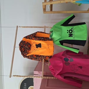 May include: Three children's hoodies hanging on a wooden rack. The hoodie on the left has a leopard print and orange fabric with a cartoon girl graphic. The middle hoodie is green with black accents and the text "N&Ouml;: No Exceptions" printed on it. The hoodie on the right is pink with a white and black graphic of balloons.