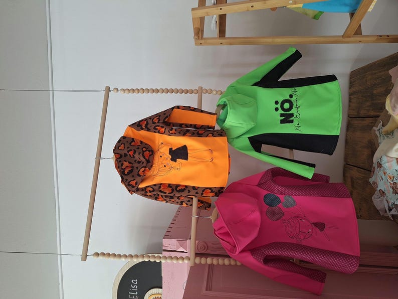 May include: Three children's hoodies hanging on a wooden rack. The hoodie on the left has a leopard print and orange fabric with a cartoon girl graphic. The middle hoodie is green with black accents and the text "N&Ouml;: No Exceptions" printed on it. The hoodie on the right is pink with a white and black graphic of balloons.