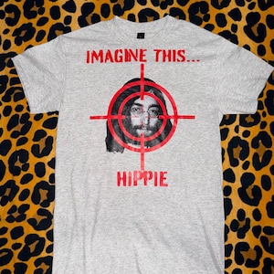 Puede incluir: Camiseta gris claro con el texto "IMAGINE THIS..." encima de un gráfico de la cara de un hombre dentro de una diana roja, con la palabra "HIPPIE" debajo. La camiseta está sobre un fondo de estampado de leopardo.
