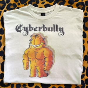 Puede incluir: Camiseta blanca con un personaje de dibujos animados Garfield musculoso, en tonos naranja y amarillo. El texto "Cyberbully" está impreso encima del personaje en una fuente gótica.