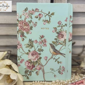 May include: A mint green notebook with a floral design featuring pink roses and a bird perched on a branch. The word "NOTES" is printed on the cover. The notebook is surrounded by decorative elements.