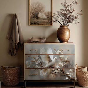 May include: A three-drawer chest of drawers with a dove and floral design. The chest has a light wood top and dark metal legs. The image also includes a framed landscape painting, a woven basket, and a large copper vase.
