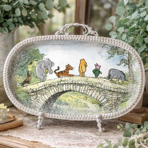 Puede incluir: Bandeja decorativa ovalada con una ilustración clásica de Winnie the Pooh y sus amigos cruzando un puente de piedra. La bandeja tiene un borde blanco con un asa decorativa y se exhibe sobre un soporte. La escena está ambientada en un bosque caprichoso.