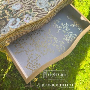 Puede incluir: Cajón abierto con un diseño floral y de cebra en la superficie superior. El interior del cajón es azul claro con detalles florales dorados. El cajón tiene molduras y herrajes dorados. El texto "re-design" y "Emporium Deluxe" son visibles.