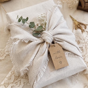 Puede incluir: Un regalo rectangular envuelto en tela de lino beige con bordes deshilachados, atado en un nudo central. Una pequeña etiqueta de papel marrón cuelga del nudo, impresa con el texto 'with love' y un símbolo de corazón. Una ramita de eucalipto y paniculata seca está insertada en el nudo.