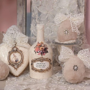 May include: A collection of decorative items, including a bottle with a floral design and text, a heart-shaped ornament with a bunny illustration, and two fluffy ornaments with lace bows. The items are in soft, neutral colors.