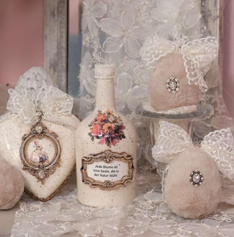 May include: A collection of decorative items, including a bottle with a floral design and text, a heart-shaped ornament with a bunny illustration, and two fluffy ornaments with lace bows. The items are in soft, neutral colors.
