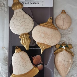 May include: A set of decorative molds for crafting, featuring various shapes of bright baubles. The molds are cream-colored with a crackle finish and gold accents. The package is 5" x 10" and includes the text "BRIGHT BAUBLES".