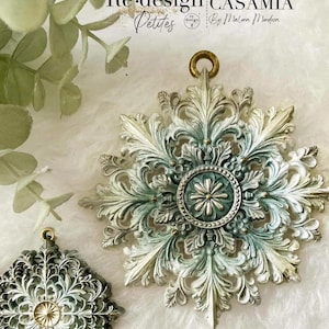 May include: Two ornate, star-shaped decorative ornaments featuring intricate acanthus leaf detailing and a central circular medallion. The larger piece displays a weathered finish in shades of sage green, cream, and metallic gold with a small hanging loop.