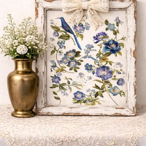 May include: A framed floral print featuring a blue bird perched among blue and purple flowers. The distressed white frame is adorned with a cream-colored lace bow. A gold vase with white flowers sits to the left.