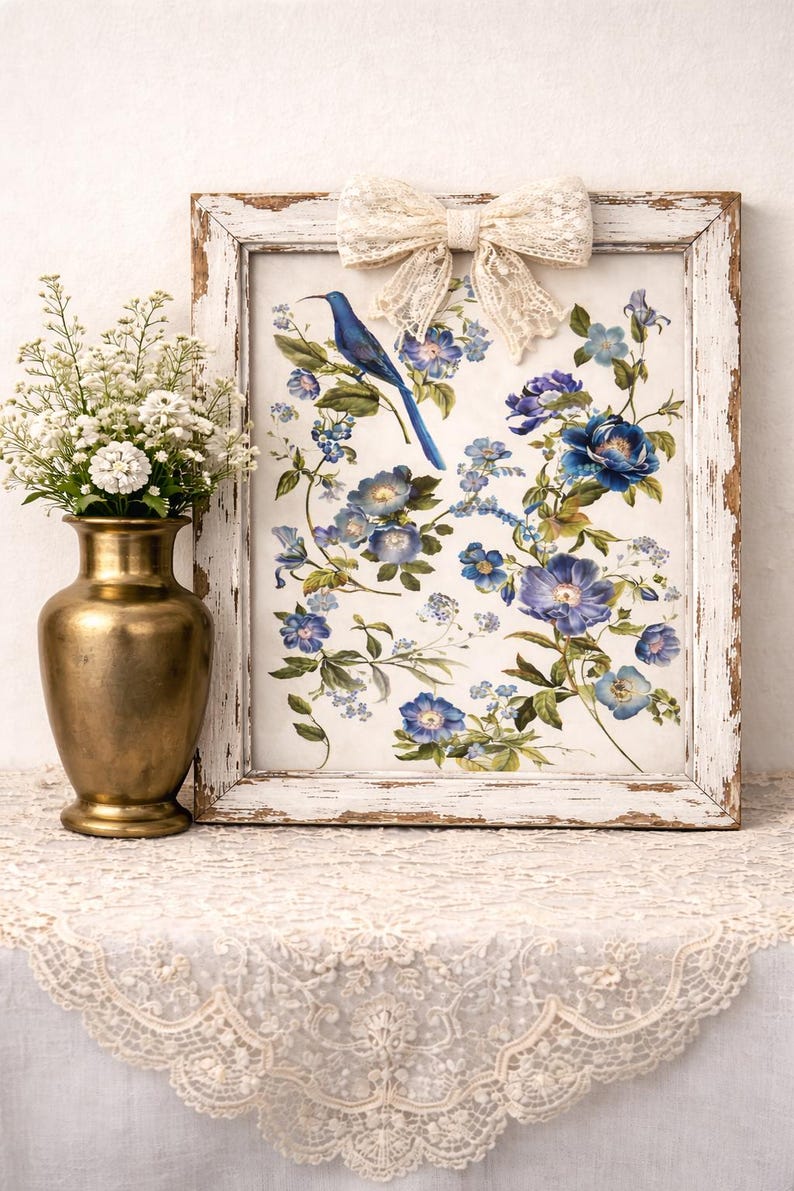 May include: A framed floral print featuring a blue bird perched among blue and purple flowers. The distressed white frame is adorned with a cream-colored lace bow. A gold vase with white flowers sits to the left.