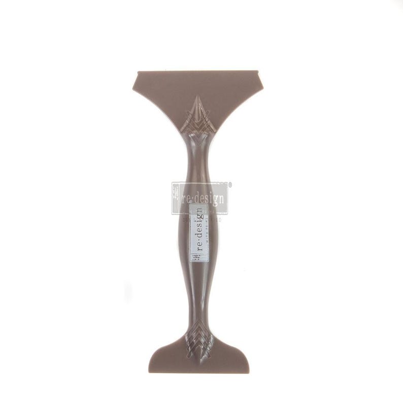 May include: A brown, decorative scraper tool with a wide, fan-shaped top and a long, slender handle. The handle features a decorative design and a label that reads "re-design". The tool is designed for craft and DIY projects.