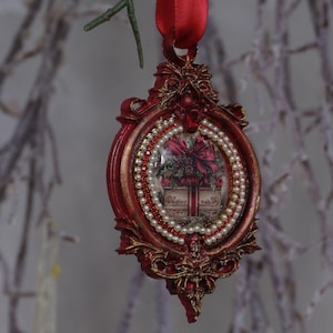 May include: Red and gold Christmas ornament with a framed design. The ornament features a picture of a gift box with a red bow, surrounded by white pearls and red beads. A red ribbon is attached for hanging.