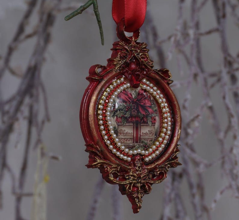 May include: Red and gold Christmas ornament with a framed design. The ornament features a picture of a gift box with a red bow, surrounded by white pearls and red beads. A red ribbon is attached for hanging.