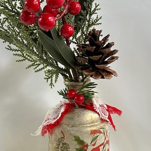 May include: A decorative Christmas-themed vase with a festive arrangement. The vase is adorned with holiday imagery and a red ribbon. The arrangement includes red berries, pine needles, and a pine cone, creating a seasonal display.