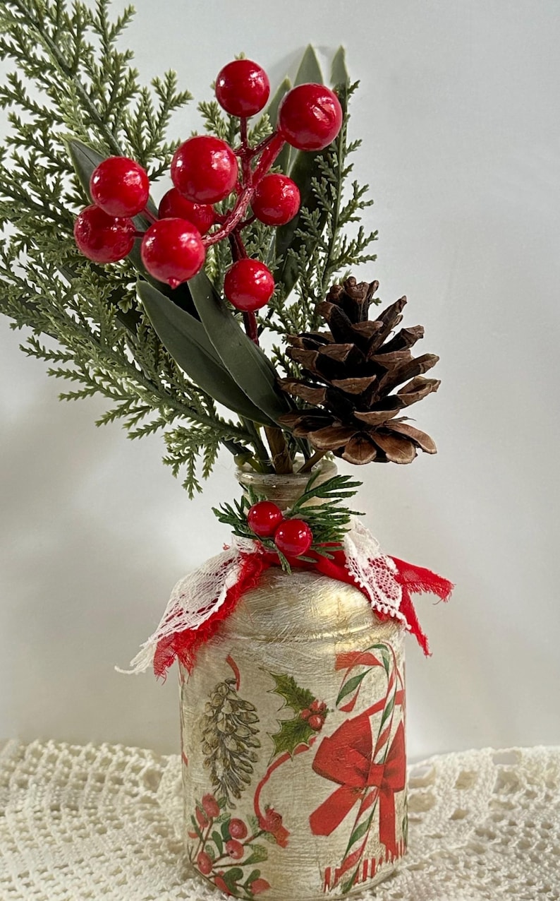 May include: A decorative Christmas-themed vase with a festive arrangement. The vase is adorned with holiday imagery and a red ribbon. The arrangement includes red berries, pine needles, and a pine cone, creating a seasonal display.