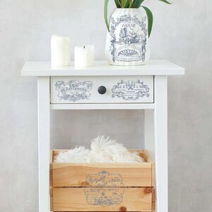 May include: A white nightstand with a drawer and a lower shelf. The drawer has decorative labels and a black knob. A wooden crate sits on the shelf, and a vase with a floral design and two candles are on top. The vase has the word "Paris" on it.