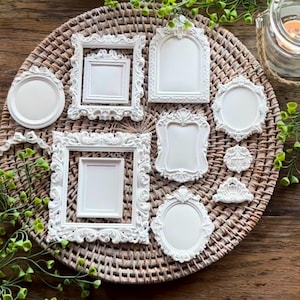 May include: A collection of white, ornate picture frames in various shapes and sizes, including round, oval, square, and arched designs. The frames are arranged on a woven, circular placemat with greenery.