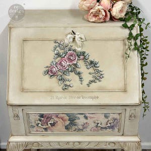 May include: An antique off-white secretary desk with floral accents. The top panel features a raised floral design with pink roses and green leaves. The drawer front has a floral pattern, and the text "21, Rue de l'Arc de Triomphe" is visible.