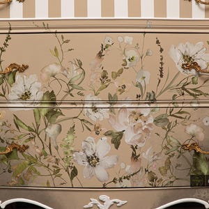 Peut inclure: Commode beige avec des œuvres d'art florales sur les tiroirs. Les fleurs sont blanches et roses avec des feuilles vertes. Le haut de la commode est rayé beige et blanc. Poignées dorées.