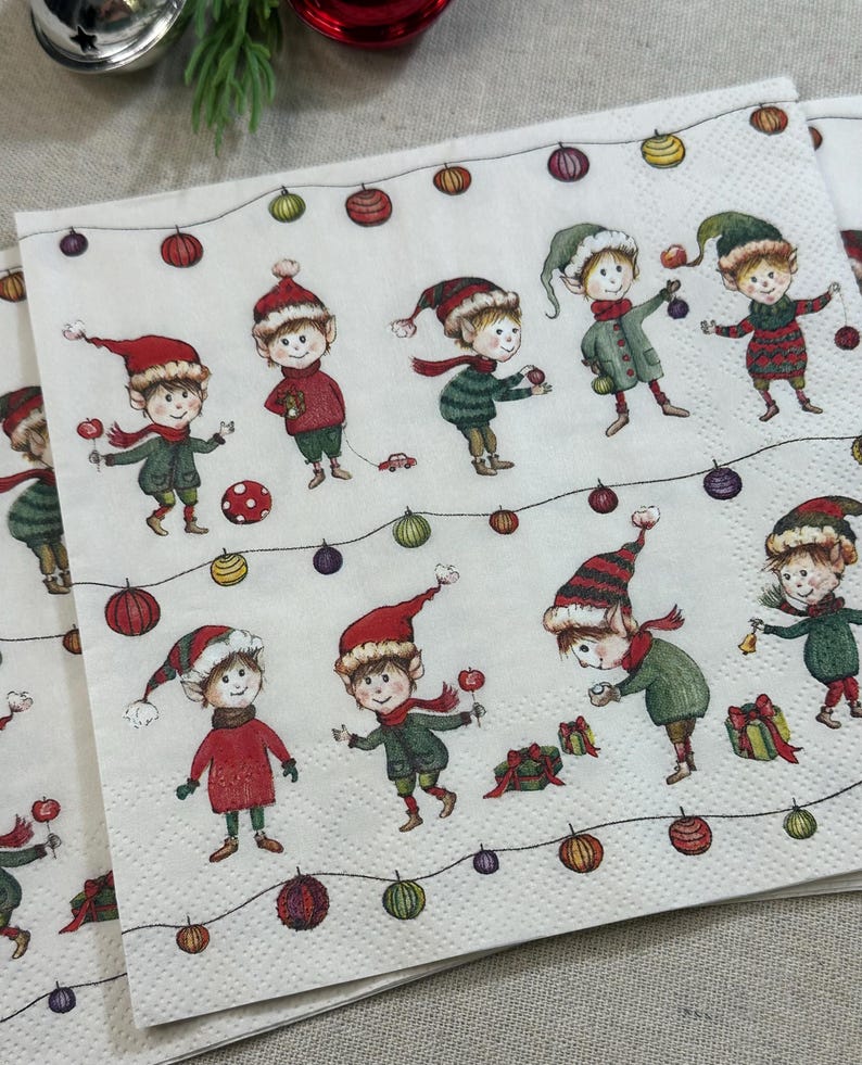 May include: White paper napkins with a repeating pattern of cartoon elves in red and green outfits, wearing hats and scarves. The elves are surrounded by colorful Christmas lights and ornaments.