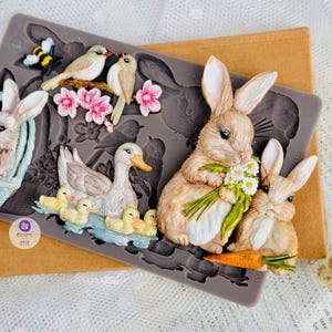 May include: A silicone mold featuring detailed animal designs. The mold includes a duck with ducklings, two rabbits, birds, a bee, and flowers. The animals are in shades of brown, yellow, and white, with pink flowers. The mold is gray.