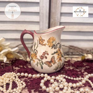 Puede incluir: Una jarra de cerámica de color crema con acabado agrietado, que presenta un asa de color rojo oscuro y un estampado decorativo de flores amarillas y una mariposa. La jarra tiene un pico curvo y un cuerpo redondeado con diseño botánico.