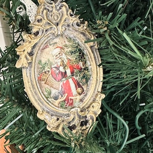 May include: Ornate Christmas ornament with a vintage design. The oval-shaped ornament features a picture of Santa Claus in a red suit, holding a basket. The frame is gold with an aged finish, and the ornament hangs from a white lace ribbon. The text "'Twas to you" is visible.