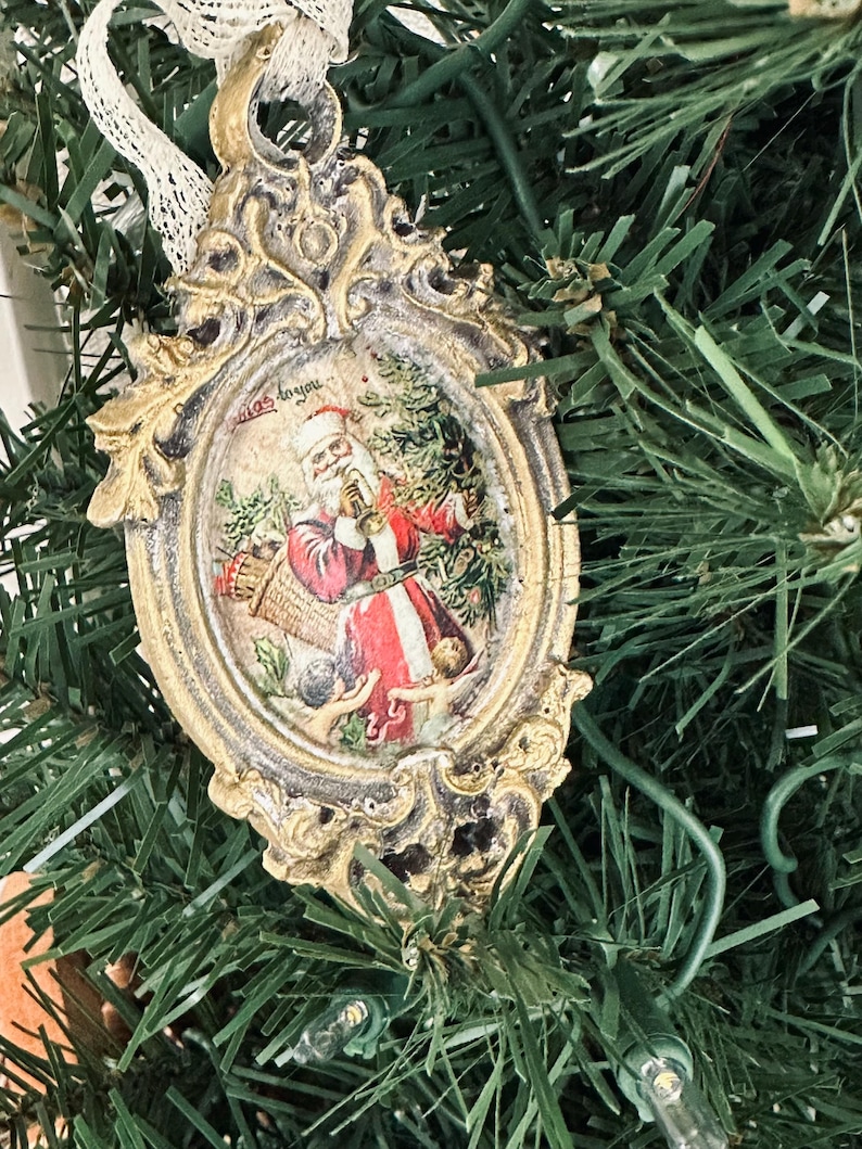 May include: Ornate Christmas ornament with a vintage design. The oval-shaped ornament features a picture of Santa Claus in a red suit, holding a basket. The frame is gold with an aged finish, and the ornament hangs from a white lace ribbon. The text "'Twas to you" is visible.