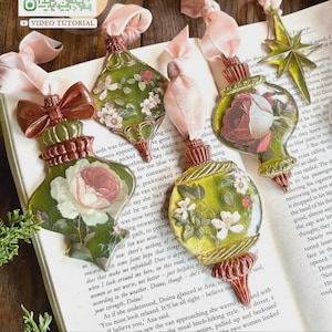 Op de afbeelding: Vier decoratieve ornamenten met bloemmotieven in groen-, roze- en wittinten, elk voorzien van een roze fluwelen lint. De ornamenten hebben metalen doppen en voetjes, inclusief een goudkleurig sterornament.