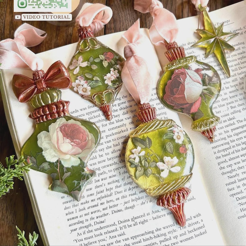 Op de afbeelding: Vier decoratieve ornamenten met bloemmotieven in groen-, roze- en wittinten, elk voorzien van een roze fluwelen lint. De ornamenten hebben metalen doppen en voetjes, inclusief een goudkleurig sterornament.