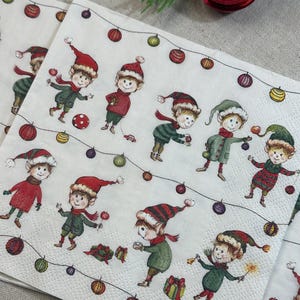 May include: White paper napkins with a festive Christmas elf design. The napkins feature multiple elves in red and green outfits, wearing hats and scarves, with string lights and ornaments. The elves are holding gifts and toys.