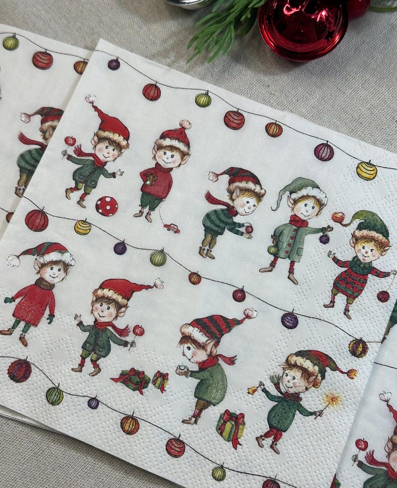 May include: White paper napkins with a festive Christmas elf design. The napkins feature multiple elves in red and green outfits, wearing hats and scarves, with string lights and ornaments. The elves are holding gifts and toys.