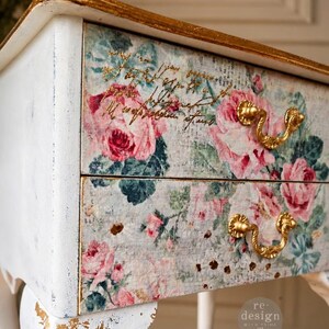 May include: A white wooden side table with two drawers featuring a floral pattern of pink roses and green leaves. The table has gold-painted edges, gold curved drawer pulls, and gold leaf accents on the legs. Text on the drawer reads re.design.