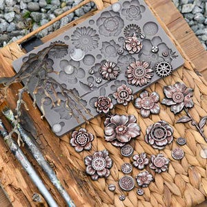 May include: A collection of steampunk-style floral embellishments and a silicone mold on a wooden surface. The embellishments are in shades of bronze and copper, with intricate gear and flower designs. Paintbrushes and a decorative branch are also present.