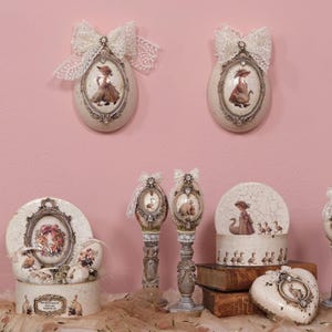 May include: Decorative egg-shaped wall hangings and decorative boxes with vintage-style illustrations of women and animals. The items are cream-colored with ornate frames, lace bows, and a heart-shaped box.
