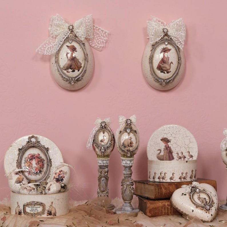 May include: Decorative egg-shaped wall hangings and decorative boxes with vintage-style illustrations of women and animals. The items are cream-colored with ornate frames, lace bows, and a heart-shaped box.
