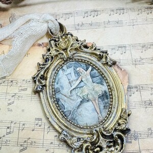 May include: Ornate gold-colored oval ornament with a picture of a ballerina in a light green dress. The frame has intricate scrollwork and is attached to a cream-colored ribbon. The background is sheet music.