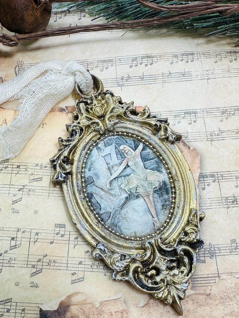 May include: Ornate gold-colored oval ornament with a picture of a ballerina in a light green dress. The frame has intricate scrollwork and is attached to a cream-colored ribbon. The background is sheet music.