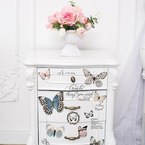 May include: A white wooden bedside table with three drawers, featuring butterfly decals and cursive text that reads dream, Create the thing you wish existed, and inspire. The table has ornate carved legs and brass-toned drawer pulls.